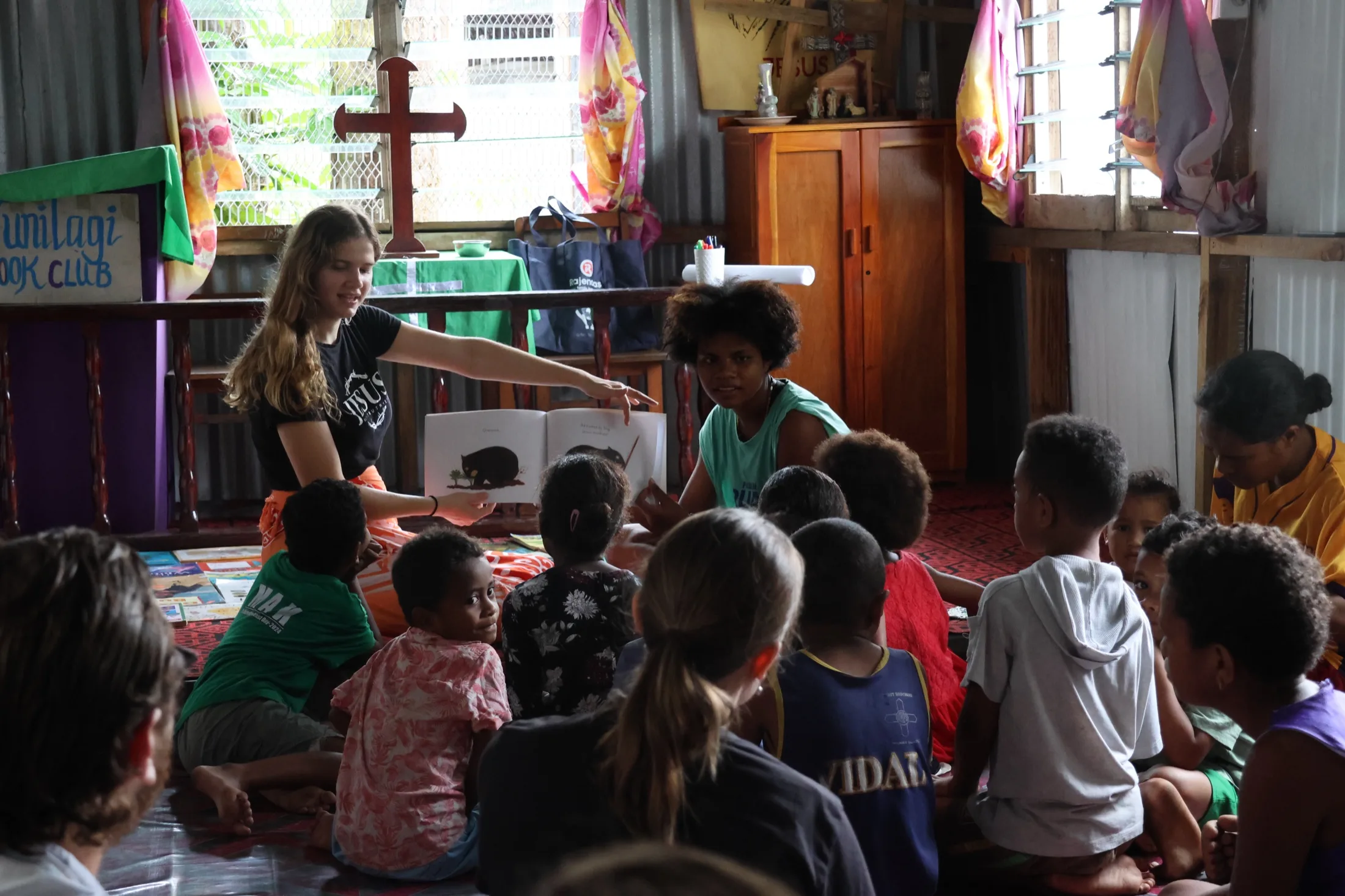 A Vunilagi volunteer reads a picture book aloud to a group of children seated together in a community hall